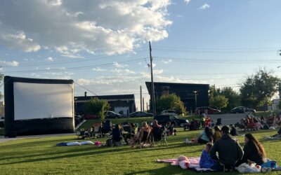 Screen on the Green: DAYTIME-CROWD A group of people sit in a grassy area, watching a movie on an outdoor screen.