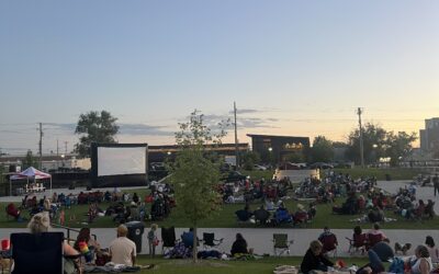 Screen on the Green: SUNSET-CROWD A group of people sit in a grassy area, watching a movie on an outdoor screen.